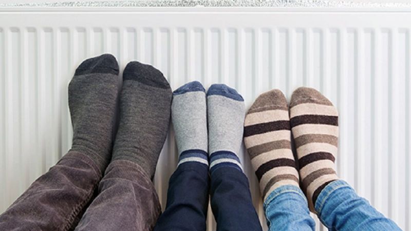 Symbolbild: Zwei Erwachsene und ein Kind wärmen ihre Füße in Socken an einem Heizkörper.