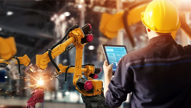 Symbolbild Künstliche Intelligenz: Arbeiter von hinten mit Laptop  vor 2 Roboterarmen in Industriehalle