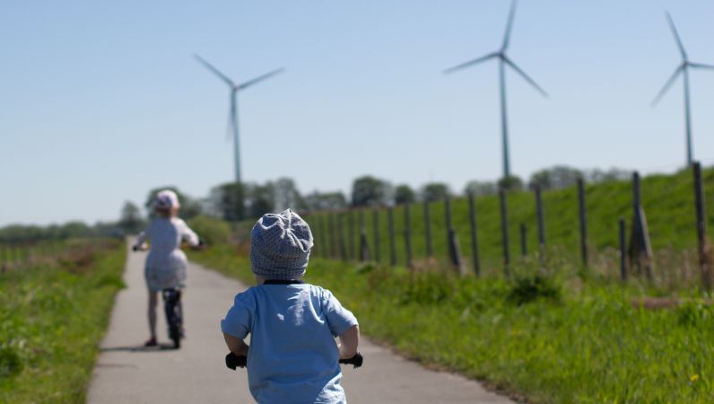 Kinder von hinten auf Fahrrädern vor Windrädern auf einem Radweg im Feld